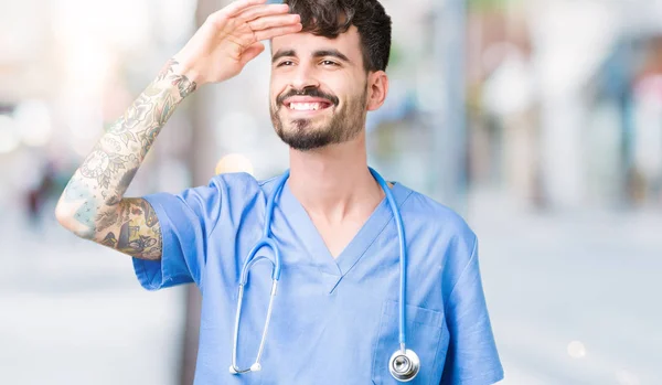 Young handsome nurse man wearing surgeon uniform over isolated background very happy and smiling looking far away with hand over head. Searching concept.