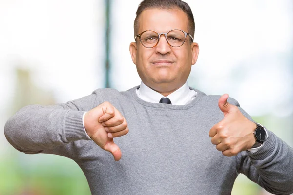 Middle age bussines arab man wearing glasses over isolated background Doing thumbs up and down, disagreement and agreement expression. Crazy conflict