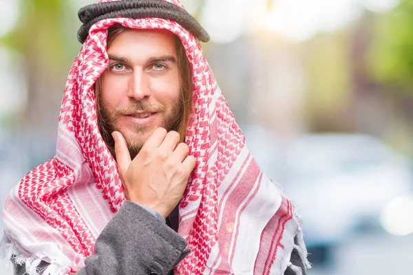 Young handsome arabian man with long hair wearing keffiyeh over isolated background looking confident at the camera with smile with crossed arms and hand raised on chin. Thinking positive.