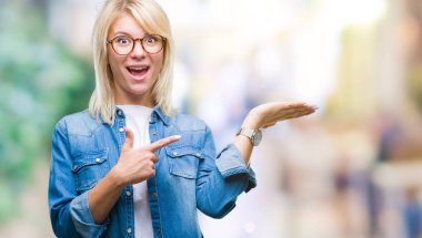 Genç güzel sarışın kadın hayrete ve el ile sunulması ve parmak ile işaret ederken kameraya gülümseyen izole arka plan üzerinde gözlük.