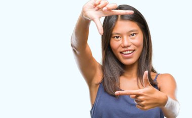 Genç Asyalı kadın elle yapma çerçeve ve parmakları ile mutlu yüz gülümseyen izole arka plan üzerinde. Yaratıcılık ve Fotoğrafçılık kavramı.