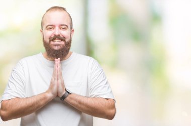 Ellerle birlikte AF emin gülümseyen için soran dua izole arka plan üzerinde rahat t-shirt giyen genç beyaz hipster adam.