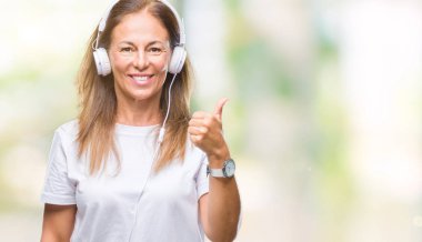 Orta yaş İspanyol kadın arıyorum ve başparmak yanında yukarıyı mutlu yüz ile gülümseyen izole arka plan üzerinde kulaklık giyen müzik dinlemek.