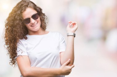 Kameraya bakarak yan el ve parmak ile işaret yüzünde kocaman bir gülümseme ile izole arka plan üzerinde güneş gözlüğü takıyor güzel esmer kıvırcık saçlı Genç kız.