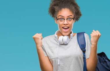 Kulaklıklar ve sırt çantası izole arka plan kutlama üzerinde giyen genç afro Amerikan öğrenci kadın şaşırttı ve başarı için kaldırdı kollarını açarak hayret ve gözleri açık. Kazanan kavramı.