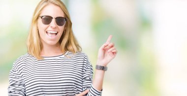 Kameraya bakarak yan el ve parmak ile işaret eden güzel genç kadın yüzünde kocaman bir gülümseme ile izole arka plan üzerinde güneş gözlüğü takıyor.