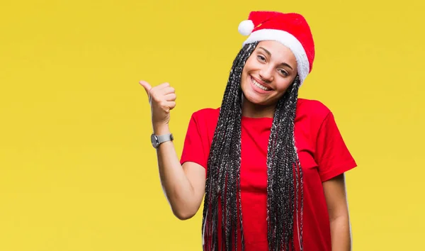 年轻的编织头发非洲裔美国女孩戴着圣诞帽在孤立的背景微笑着与快乐的脸看 用拇指指着一边 — 图库照片