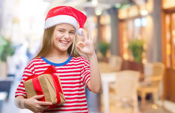 優秀なシンボルの指で サインをして孤立した背景にクリスマスの帽子 持株ギフトを着た美しい少女 — ストック写真