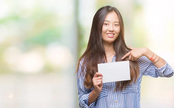 Young asian woman holding blank card over isolated background with surprise face pointing finger to himself