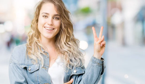 Mutlu yüz zafer işareti yaparken kameraya göz kırpıyor ile gülümseyen izole arka plan üzerinde kot ceket giyen güzel genç sarışın kadın. İki numaralı.