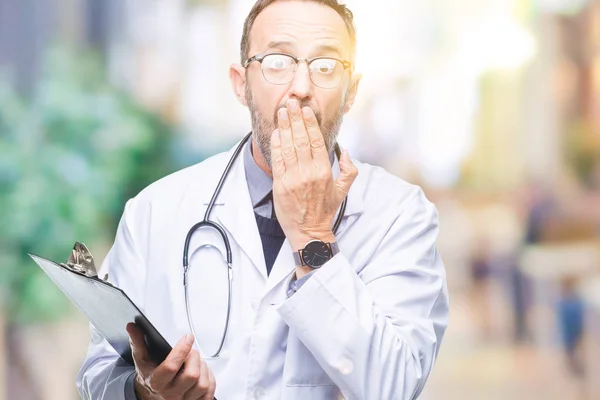 Middle age senior hoary doctor man holding clipboard over isolated background cover mouth with hand shocked with shame for mistake, expression of fear, scared in silence, secret concept