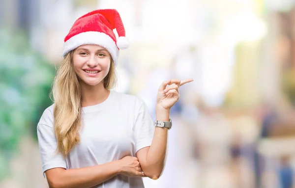 若い金髪美人の顔に大きな笑みを浮かべて隔離された背景にクリスマスの帽子を着てカメラを見て側の手と指で指す — ストック写真