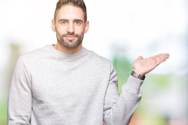 Neşeli sunulması ve kameraya bakarak avucunun ile işaret gülümseyen izole arka plan üzerinde sweatshirt giyen genç yakışıklı adam.