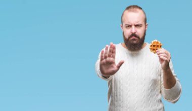 Genç beyaz hipster man açık eliyle dur işareti ile ciddi ve kendine güvenen ifade, savunma jest yapıyor izole arka plan üzerinde tatlı waffle yemek