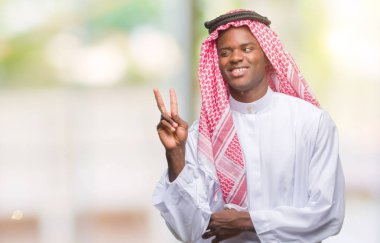 Mutlu yüz zafer işareti yaparken kameraya göz kırpıyor ile gülümseyen izole arka plan üzerinde geleneksel keffiyeh giyen genç Arapça Afrika adam. İki numaralı.