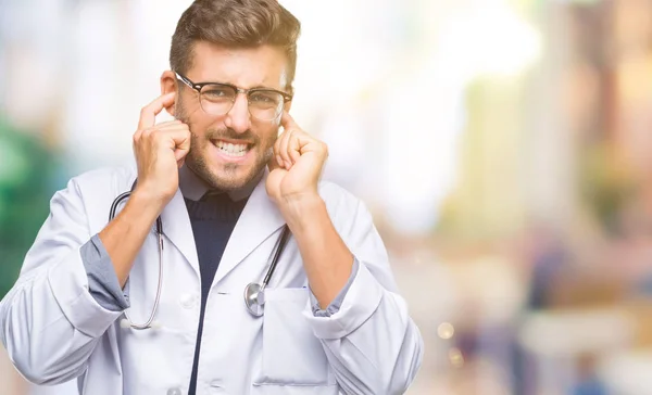 Young handsome doctor man over isolated background covering ears with fingers with annoyed expression for the noise of loud music. Deaf concept.