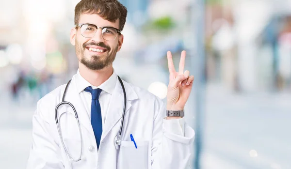 年轻的医生穿在孤立的背景上的医院外套微笑着 高兴的脸对着镜头眨眼做胜利的标志 — 图库照片