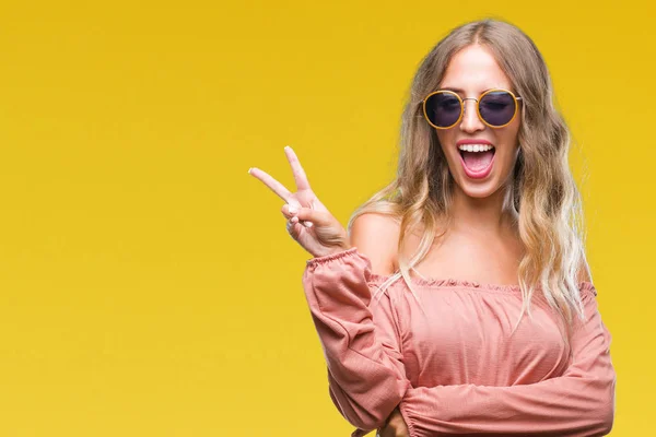 Beautiful young blonde woman wearing retro sunglasses over isolated background smiling with happy face winking at the camera doing victory sign. Number two.