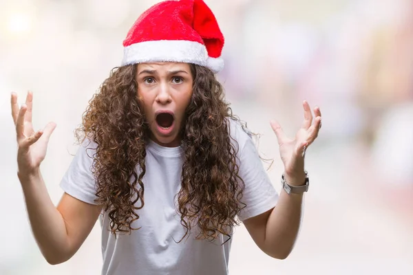 年轻的黑发女孩戴着圣诞帽在孤立的背景疯狂和疯狂的喊声和大喊大叫与侵略性的表情和手臂提出 挫折的概念 — 图库照片