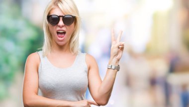 Genç güzel sarışın kadın mutlu yüz zafer işareti yaparken kameraya göz kırpıyor ile gülümseyen izole arka plan üzerinde güneş gözlüğü takıyor. İki numaralı.