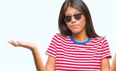 Genç Asyalı kadın izole arka plan clueless ve şaşkın bir ifadeyle kollar ve eller kaldırdı üzerinde güneş gözlüğü takıyor. Şüphe kavramı.