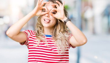 Güzel sarışın genç kadın dilini, parmaklarının arasından baktığını yapışmasını dürbün gibi ok jest yapıyor izole arka plan üzerinde. Deli ifade.