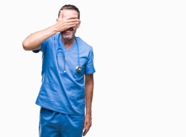 Orta yaş ağarmış kıdemli Doktor adam gülümsüyor ve gözleri için sürpriz kapsayan yüz el ile gülüyor izole arka plan üzerinde tıbbi üniforma. Kör kavramı.