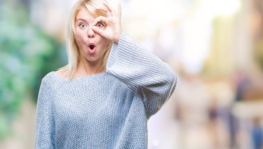 Sürpriz yüz, göz parmaklarının arasından seyir ile ok jest yapıyor izole arka plan üzerinde kış kazak giyen genç güzel sarışın kadın şok. İmansız ifade.