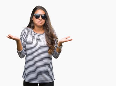 Genç güzel Arap kadın üzerinde güneş gözlüğü takıyor arka plan clueless ve şaşkın bir ifadeyle kollar ve eller kaldırdı izole. Şüphe kavramı.