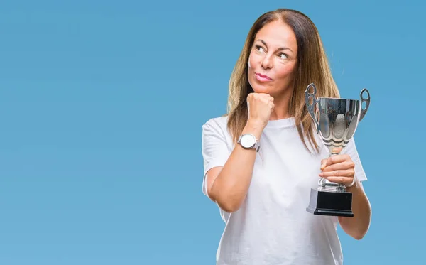Middle age hispanic winner woman celebrating award holding trophy over isolated background serious face thinking about question, very confused idea