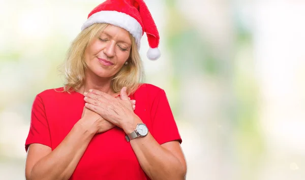 Middle age blonde woman wearing christmas hat over isolated background smiling with hands on chest with closed eyes and grateful gesture on face. Health concept.