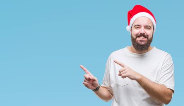 Gülümseyen ve iki elleri ve parmakları ile tarafına işaret kamera bakarak izole arka plan üzerinde Noel şapka giyen genç beyaz hipster adam.