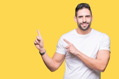 Gülümseyen ve iki elleri ve parmakları ile tarafına işaret kamera bakarak izole arka plan üzerinde rahat beyaz t-shirt giyen genç adam.