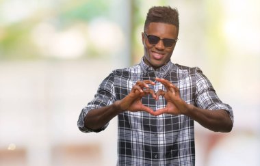 Genç Afro-Amerikan adam aşık kalp simgesi ve elleriyle şekil gösteren gülümseyen izole arka plan üzerinde güneş gözlüğü takıyor. Romantik kavramı.