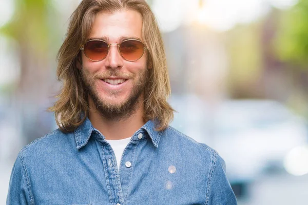Genç yakışıklı adam uzun saçlı yüzünde mutlu ve güzel bir gülümseme ile izole arka plan üzerinde güneş gözlüğü takıyor. Şanslı kişi.