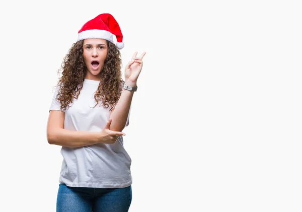 年轻的黑发女孩戴着圣诞帽在孤立的背景微笑着 幸福的脸在相机眨眼做胜利的标志 — 图库照片