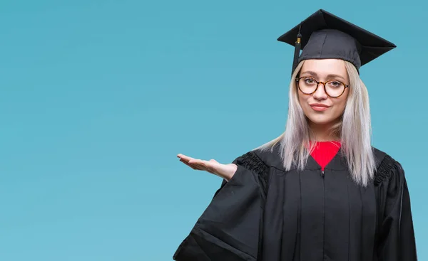 Young blonde woman wearing graduate uniform over isolated background smiling cheerful presenting and pointing with palm of hand looking at the camera.