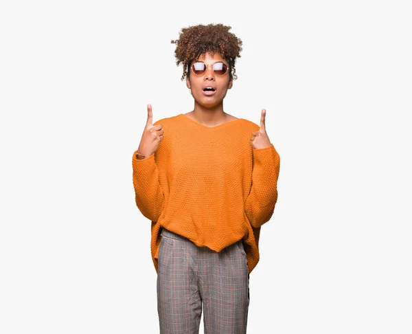 美丽的年轻非洲裔美国妇女戴着太阳镜在孤立的背景惊讶和惊讶地抬头 用手指和举起的手臂指指点点 — 图库照片