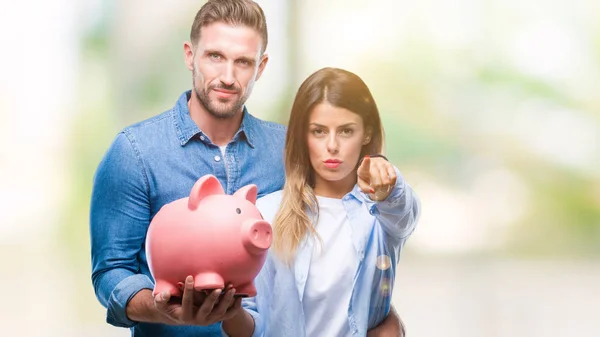 Young couple in love holding piggy bank over isolated background pointing with finger to the camera and to you, hand sign, positive and confident gesture from the front