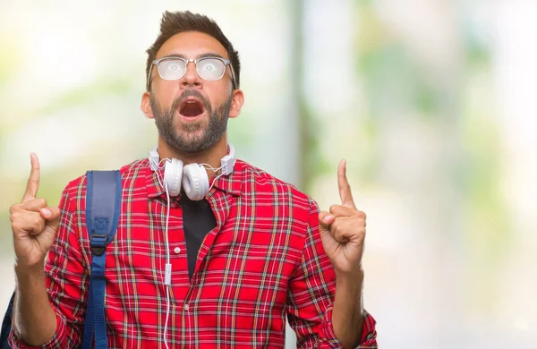 Kulaklıklar ve sırt çantası izole arka plan üzerinde giyen yetişkin İspanyol öğrenci adam hayret ve yukarıya bakıp parmakları ile işaret şaşırttı ve silah kaldırdı.