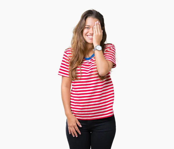 年轻美丽的黑发女人穿着条纹 T恤在孤立的背景覆盖一只眼睛与自信的微笑在脸上和惊喜的情感 — 图库照片