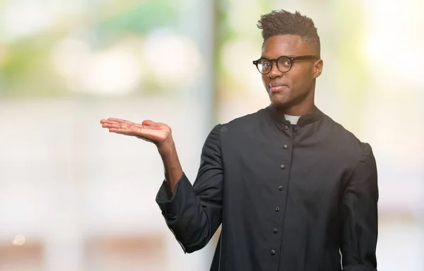 Young african american priest man over isolated background smiling cheerful presenting and pointing with palm of hand looking at the camera.