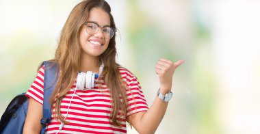 Kulaklıklar ve sırt çantası arıyorum ve başparmak yanında işaret mutlu yüz ile gülümseyen izole arka plan üzerinde giyen genç güzel esmer öğrenci kadın.