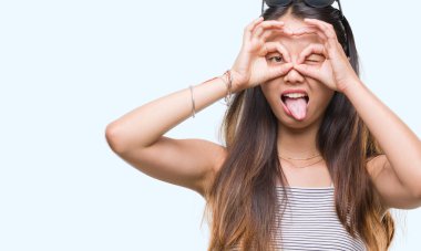 Genç Asyalı kadın dilini, parmaklarının arasından baktığını yapışmasını dürbün gibi ok jest yapıyor izole arka plan üzerinde güneş gözlüğü takıyor. Deli ifade.