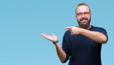 Genç beyaz hipster adam hayret ve el ile sunulması ve parmak ile işaret ederken kameraya gülümseyen izole arka plan üzerinde güneş gözlüğü takıyor.