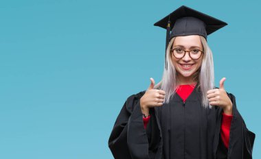 Sarışın genç kadın üzerinde el ile pozitif jest yapıyor izole arka plan başarı işareti lisansüstü üniforması başparmak gülümseyen ve mutlu. Neşeli bir ifadeyle, kazanan jest kameraya bakıyor.