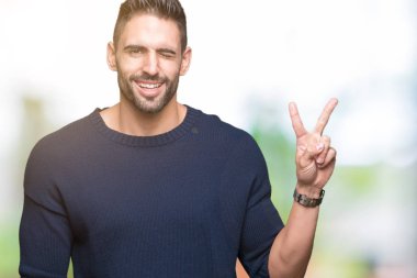 Mutlu yüz zafer işareti yaparken kameraya göz kırpıyor ile gülümseyen izole arka plan üzerinde kazak giyen genç yakışıklı adam. İki numaralı.