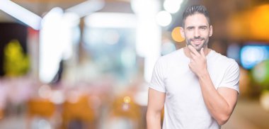 Emin çapraz kollar ve çeneme kaldırdı el ile gülümseme ile kameraya bakarak izole arka plan üzerinde rahat beyaz t-shirt giyen genç adam. Pozitif düşünce.