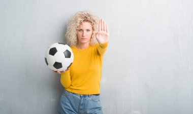 Sarışın genç kadın grunge üzerinde gri arka plan açık eliyle dur işareti ile ciddi ve kendine güvenen ifade, savunma jest yapıyor futbol futbol topu tutan