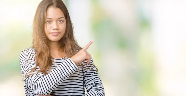 年轻美丽的黑发女人穿着条纹毛衣在孤立的背景指向用手指的一侧显示广告 严重和平静的脸 — 图库照片